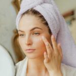 Woman with head towel applying moisturizer in front of mirror indoors., tags: haut und - pexel