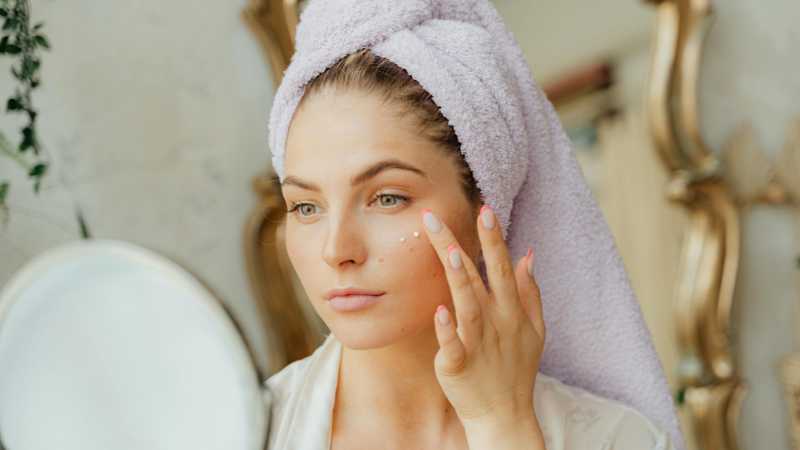 Woman with head towel applying moisturizer in front of mirror indoors., tags: haut und - pexel